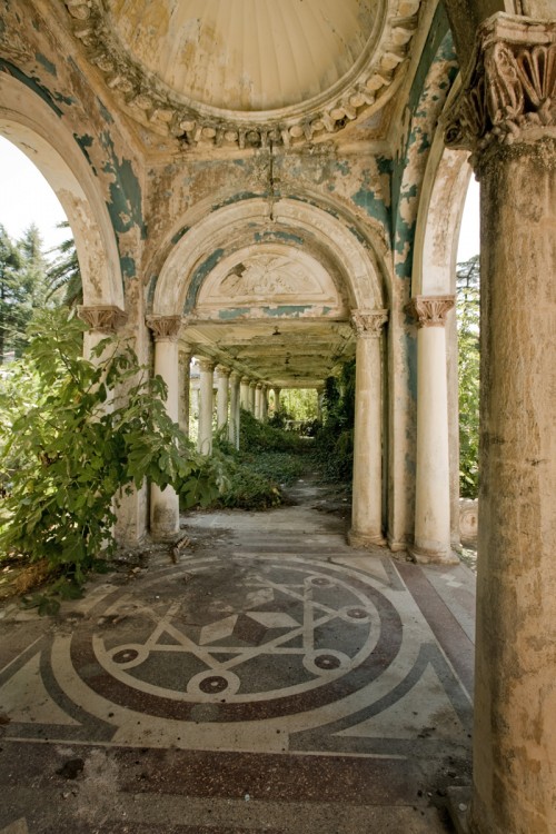 Abandoned railway station in Abkhazia A photo of a walkway in an abandoned railway station. The architecture is ornate but deteriorating, and plants have started to overgrow parts of the structure.