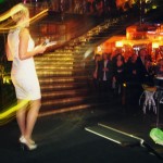 Emily Jade O'Keeffe hosts A slim white woman wearing an ivory sequinned dress stands on the stage on the left, the crowd is to the right.