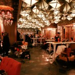 Backstage, designers, models and stylists aflurry Photo of a long room with white lanterns and dusty pink light fittings and walls, make up artists tending to models on the left and designers sitting on the right.