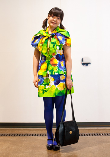 From Hel Looks A street style photo of a young woman wearing a brightly printed dress of acid green, yellow, purple and blue wearing blue tights and mary jane style shoes, carrying a big black shoulder bag.