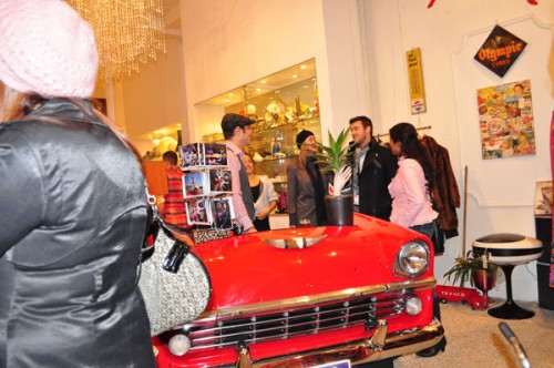 Opening night of Vintage Garage Photo of a shop with the hood of a red car in the center of the room, people crowded around talking to each other.