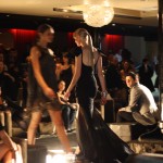 MLY BLACK LABEL Two models pass each other on the runway, the model in focus is wearing a black sleeveless tight dress with a flowing black mesh train.