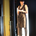 Our charming gorgeous host A woman stands behind a microphone reading from prompt cards. She wears a black leather dress, black leggings and black sandals.