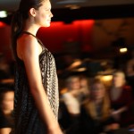 MLY BLACK LABEL Profile of a model walking down the runway in a floaty dress in a dark small pattern.
