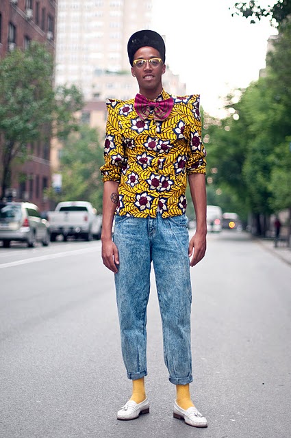 From SWAGGER 360 A street style photo of a young slim brown skinned man wearing a bright yellow print shirt with pointy shoulders, big pink bow tie, pegged and too short stone wash jeans with yellow socks and cream loafers. He also wears a cap tilted back on his head and yellow rimmed glasses.