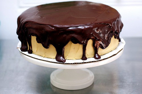 A chocolate peanut butter cake sitting on a cake dish - it looks delectable, with thick dark chocolate icing dripping down the sides of the buttery yellow cake.