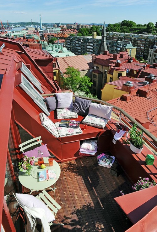 A red-painted deck overlooking densly crowded rooftops in a city. Cushions, blankets, magazines and plants are scattered on the horizontal surfaces on the deck. 