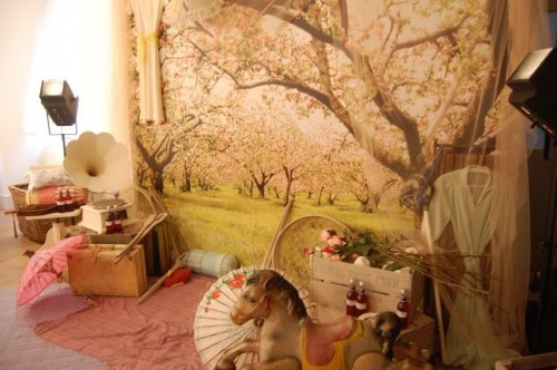 A photo of a room with a giant picture of a cherry blossom tree (I think!) on the wall and lots of whimsical things strewn about on the floor including: parasols, rocking horses, old suitcases and an old-fashioned record player with a trumpet device (can't remember the name of this!)