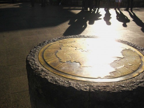 A photo of a bronze plate in a column with a map envgraved on it. The sun is long and there are long shadows of four people in the background.