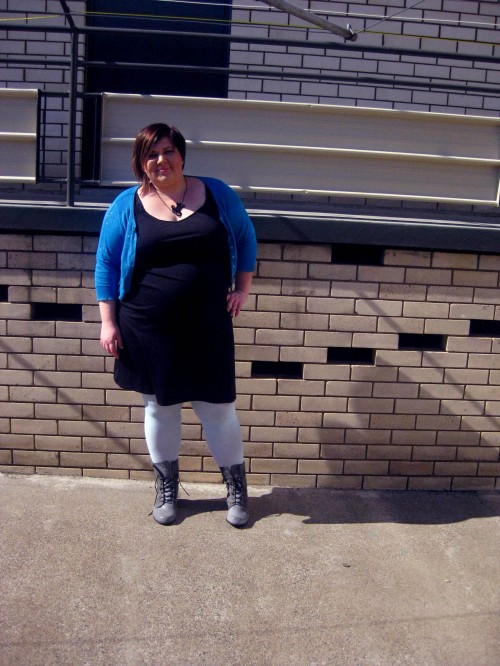 Photo of me posing for an outfit photo, wearing a black dress, pale blue tights, grey boots and a blue sparkly cardigan.