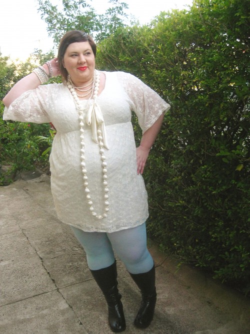 Outfit photo of me wearing a cream lace dress with big sleeves that are elbow length, mint coloured tights, black boots, and lots of cream and pearl necklaces. One hand is on my hip, the other behind my head.