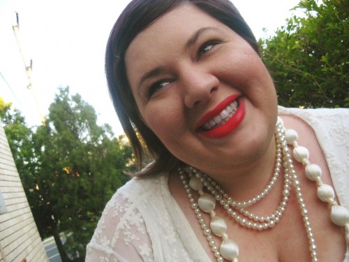 Photo of me looking off to the left, wearing bright orange/ red lipstick and lots of pearl and cream necklaces.