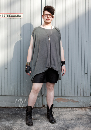 Street fashion photo of a tall white man with a big messy pomp hairstyle, turtoise shell glasses, baggy grey tshirt over black shorts and boots. He's wearing a black glove on his right hand and a black wristband on his left hand.