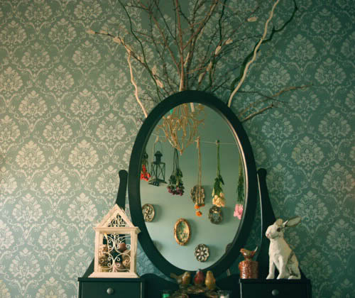 Photo of a black framed oval mirror on a bedroom vanity against a pretty teal patterned wallpaper. On the vanity include lots of trinkets including a rabbit, some birds and twigs. In the reflection of the mirror are lots of necklaces hung on a teal painted wall and some framed objects.