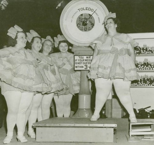 A vintage photograph from the 40s (I think!?) of four fat women looking on as another fat woman steps onto a giant scale. They're all wearing matching baby-doll dresses and bows in their hair.