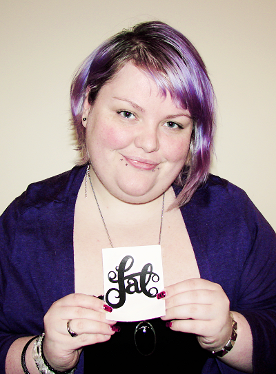 A fat pale skinned woman with short shiny mauve hair holding a sticker that says "fat" in curvy typography.