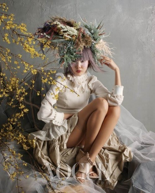 Photo of a beautiful young Asian woman wearing a white dress with feathery plumes in her lilac coloured hair. She's sitting on a mountain of coffee coloured and white material, some tulle.