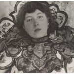 Black and white photo of a young pale skinned woman with chin length hair wearing an elaborately layered and embroidered neckpiece with scalloped edges. A rose sits on her left shoulder, she stares at the camera with an unsmilling, but not unkind, face