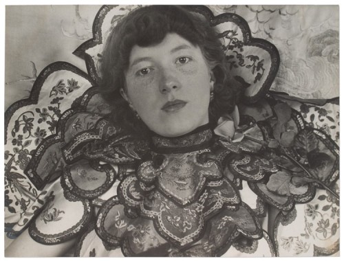 Black and white photo of a young pale skinned woman with chin length hair wearing an elaborately layered and embroidered neckpiece (or dress?) with scalloped edges. A rose sits on her left shoulder, she stares at the camera with an unsmilling, but not unkind, face