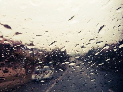 Close up photo of raindrops on a car window. Through the window you can see a suburban road.