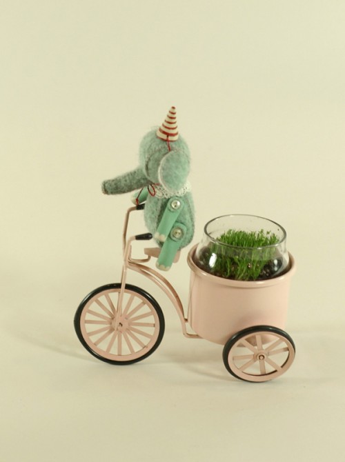 Photo of a miniature elephant in a party hat on a tricycle that holds a tiny glass jar full of grass. 