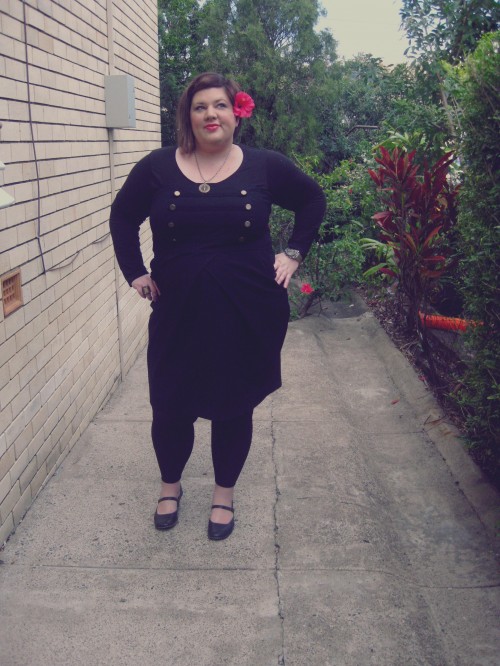 Outfit photo of me wearing a black dress with brass military style buttons at the bust, a long sleeve top under neath it and black leggings with black mary janes and a hibiscus flower behind my ear.