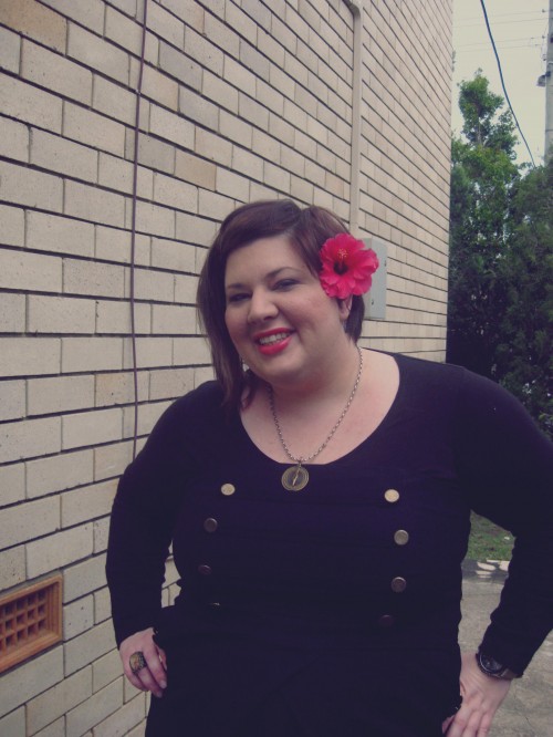 Photo of me from the waist up, wearing a black dress with military style brass buttons at the bust and a red hibiscus behind my ear. 