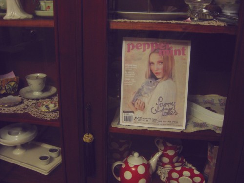 Photo of a display cabinet with Peppermint magazine inside it along with other things like tea cups and saucers and candles. The magazine has a young blonde woman holding a rabbit on the cover, and my lettering in the lower right says "furry tales".