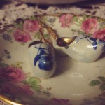 Macro shot of a pair of tiny white porcelain clogs with a blue windmill on each toe. They sit in a small condiment bowl that is bordered with a beautiful floral pattern with large pink roses.