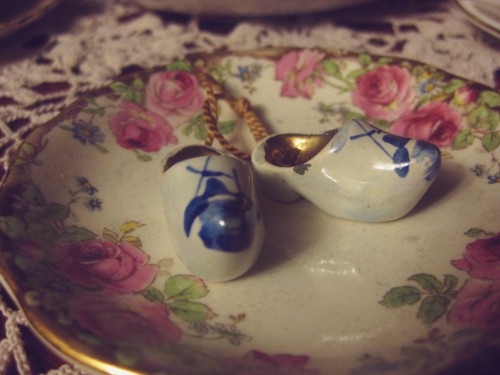 Macro shot of a pair of tiny white porcelain clogs with a blue windmill on each toe. They sit in a small condiment bowl that is bordered with a beautiful floral pattern with large pink roses.