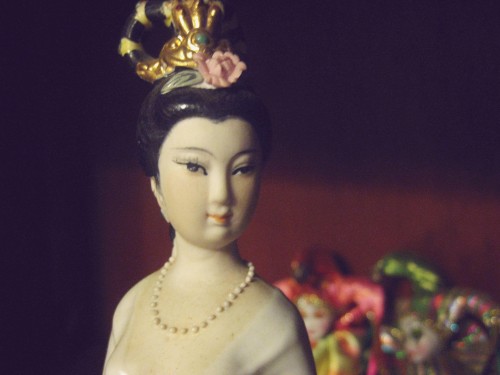 Macro photo of the face of a Chinese woman statuette, her hair is up and a  fancy ornament adorns her head. She wears a string of pearls and a white gown with a low neckline.