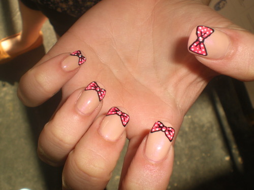 Photograph of a pale skinned hand displaying a manicure where the nail is painted beige (to match the skin tone) and a pink polka-dotted bow sits on the top of each nail. 