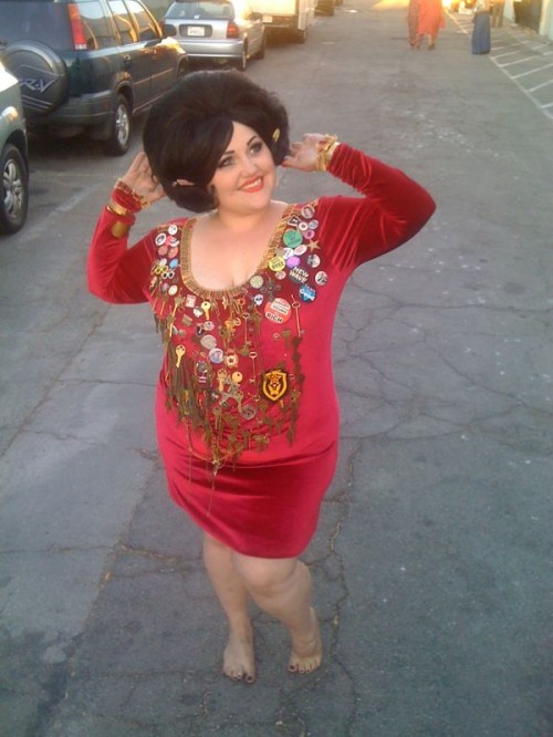 Photograph of a pale skinned fat woman with a short brown bouffant bob, wearing a cherry coloured velvet dress with lots of stuff pinned around the neckline and around the front.