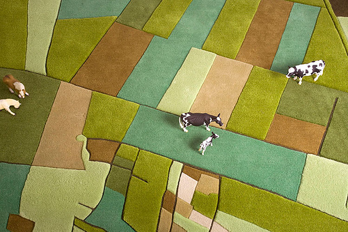 A photo of a rug that is coloured and divided like farming land as if you were viewing it from a plane. A few miniature cows are placed comically on the rug.
