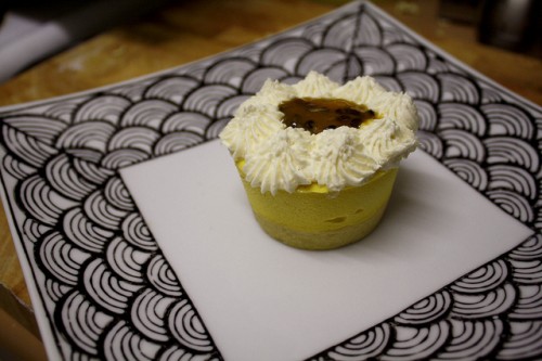 Photo of a single serving of vegan passionfruit cheesecake on a big square plate with black "fishscale" pattern bordering it.