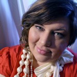 A close up photo of me wearing a red dress and a number of big pearly bead necklaces. I'm grinning/ smirking and my hair is a little bit wavy.