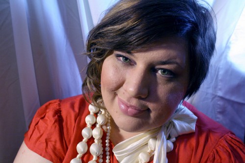 A close up photo of me wearing a red dress and a number of big pearly bead necklaces. I'm grinning/ smirking and my hair is a little bit wavy.