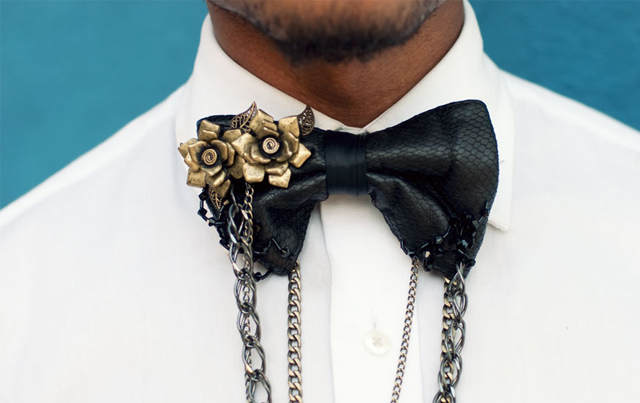 Photo of a black leather look bow tie on a brown skinned man wearing a white shirt. The tie has brass flowers in the left corner and has chains of different widths draping from it. 