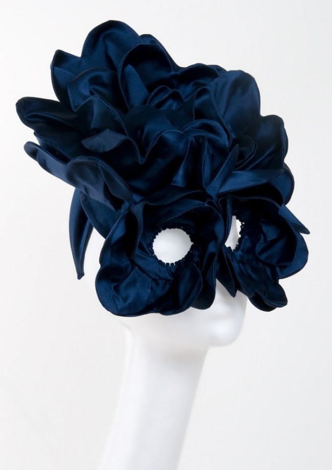 Photo of a huge black silk headpiece on a white model head, it's all ruffly and circles the eyes like a mask.