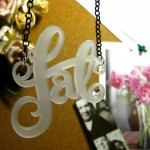 Close up shot of my fat necklace cut in a frosted white acrylic. It's hanging in front of my pin board which has some fake flowers, photobooth photos and a photo of a nice floral arrangement pinned to it.