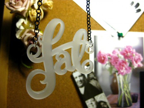 Close up shot of my fat necklace cut in a frosted white acrylic. It's hanging in front of my pin board which has some fake flowers, photobooth photos and a photo of a nice floral arrangement pinned to it.
