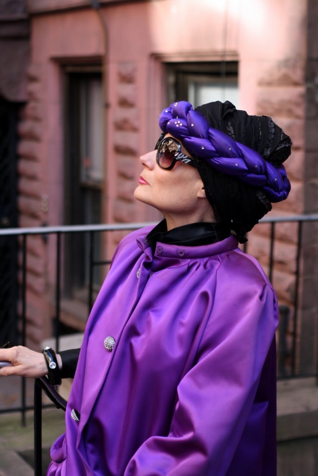 Photo of an older woman with pale skin wearing a black hair wrap with a purple plaited headband around it, as well as a magnificent purple coloured jacket. She wears dark sunglasses and is gazing off into the distance in profile.