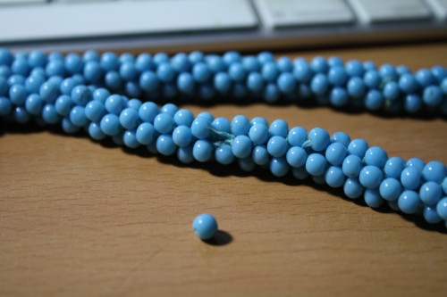 A macro photo of a multistrand turquoise beaded necklace with a broken thread poking out of the tightly beaded rope. The offending single bead sits in front of the necklace.