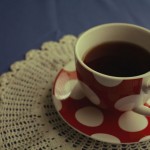 Photo of a red teacup full of black tea with big white polkadots sitting on a cream doily, which is sitting on a blue tablecloth.