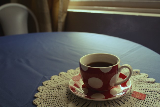 Photo of a red teacup with big white polkadots filled with black tea. The teabag is wound around the handle, and the teacup sits on a cream doily which sits on a blue tablecloth.