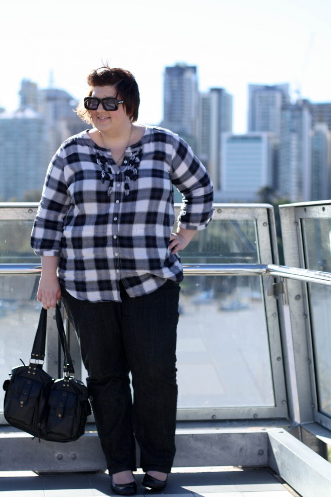 Outfit photo of me wearing a black checked tunic top with small ruffles down the button front, black jeans and round toe black wedges with a black bag. Behind me is a blurry backdrop of the CBD of Brisbane.