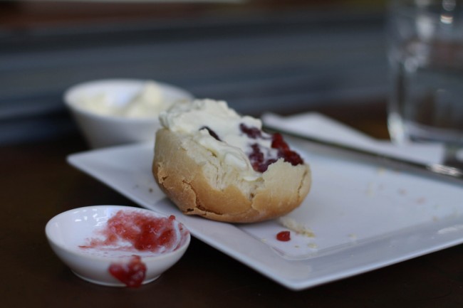 Photo of half a scone with jam and cream on a plate. An empty dish of jam sits to the side.
