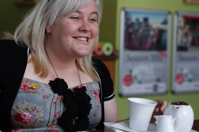 Photo of a pale skinned young woman with almost white shoulder length hair talking and smiling. There's a tea set at her hand.