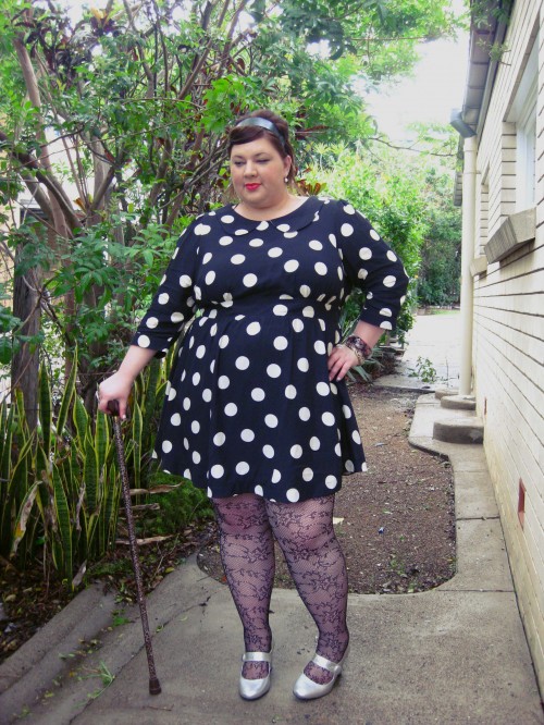 Outfit photo of me in a black dress with ivory polka dots. It has a little Peter Pan collar, and the skirt hits mid thigh. I'm wearing lace fishnet tights with silver mary jane heels, and I have my leopard print walking stick.