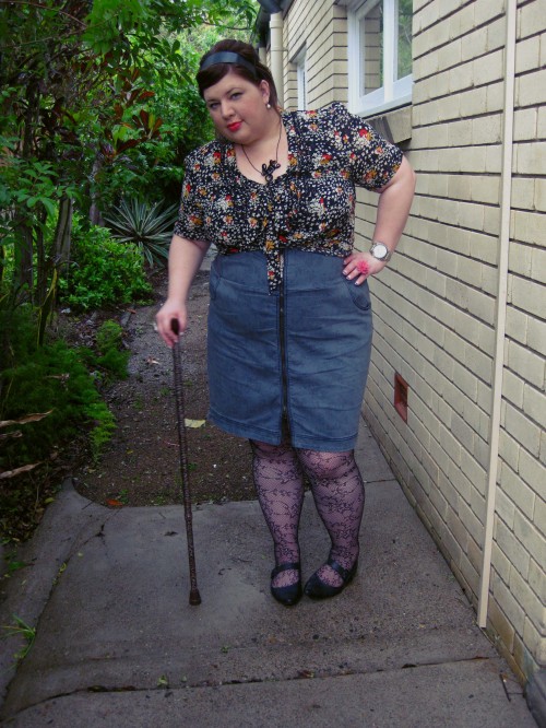 Outfit photo of me wearing a dark patterned blouse and a grey high waised pencil skirt with a zip up the front. I'm wearing lace fishnet stockings and flat pointy mary jane shoes. I'm leaning on a leopard printed walking stick.